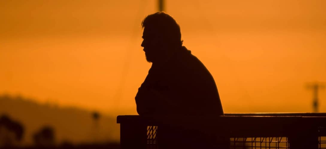 Silhouette of man with beard at sunset leaning on fence looking out at view