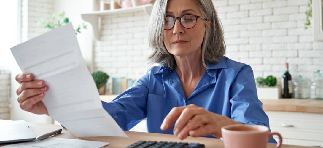 Mature woman holding paper bill using calculator managing finances at home